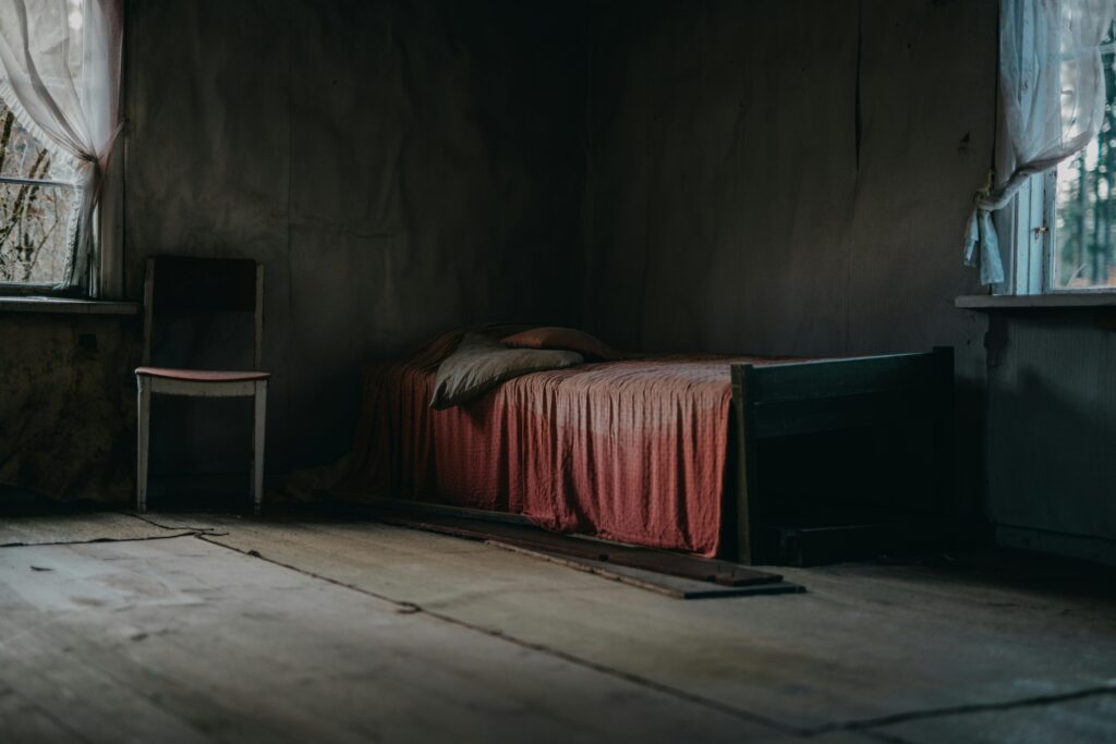 The Whisper Under the Bed Moody, textured room with an old bed and wooden chair showcasing abandonment.