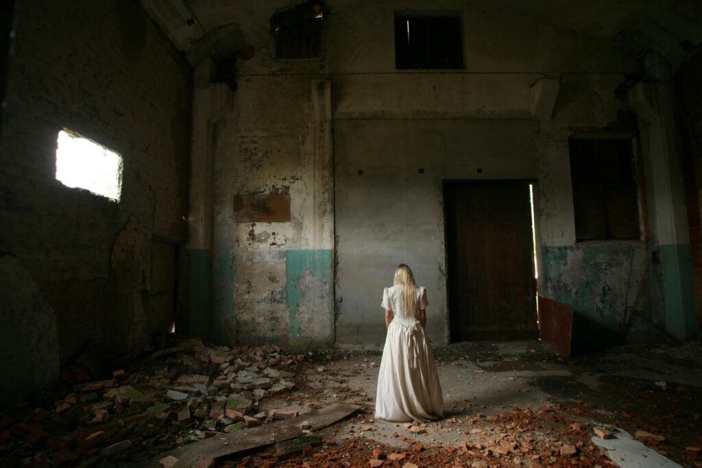 A woman in white stands in an eerie, abandoned building with decayed walls.