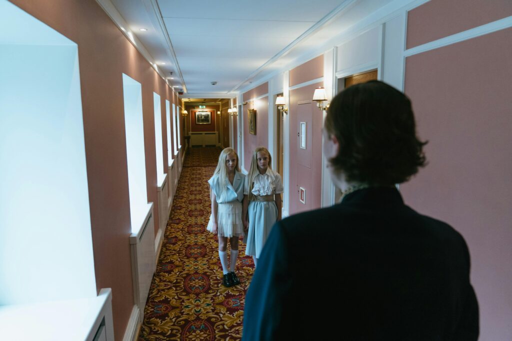 Two girls stand in a vintage hotel corridor, facing a man in the distance.