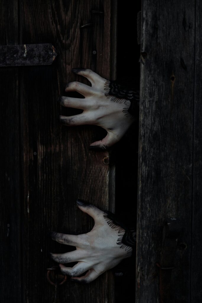 Eerie white hands with black nails reach through a dark wooden door, perfect for Halloween themes.